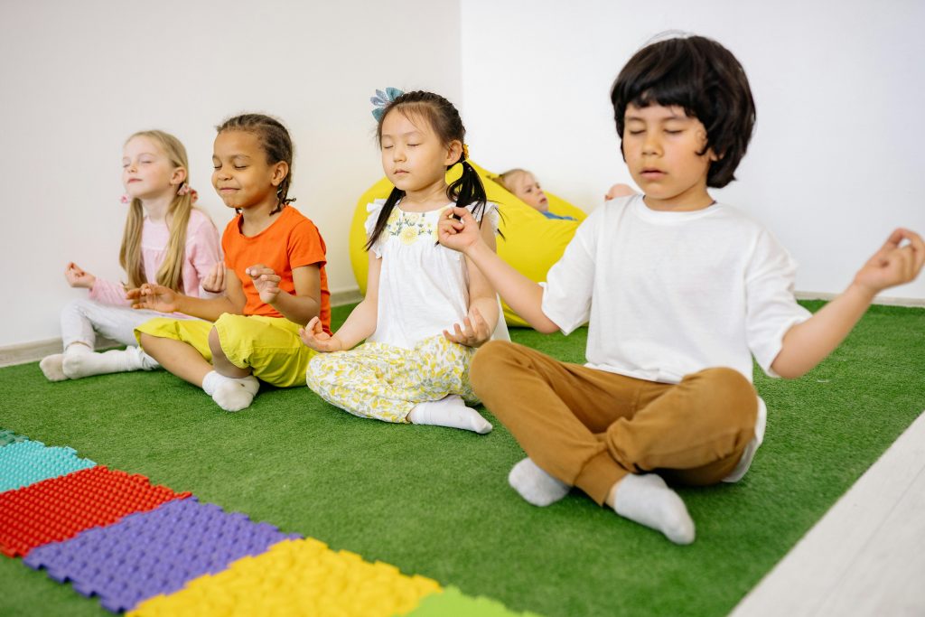 four boys and girls sitting on a turf mat crossing their legs with their eyes closed in front of colorful foam mat