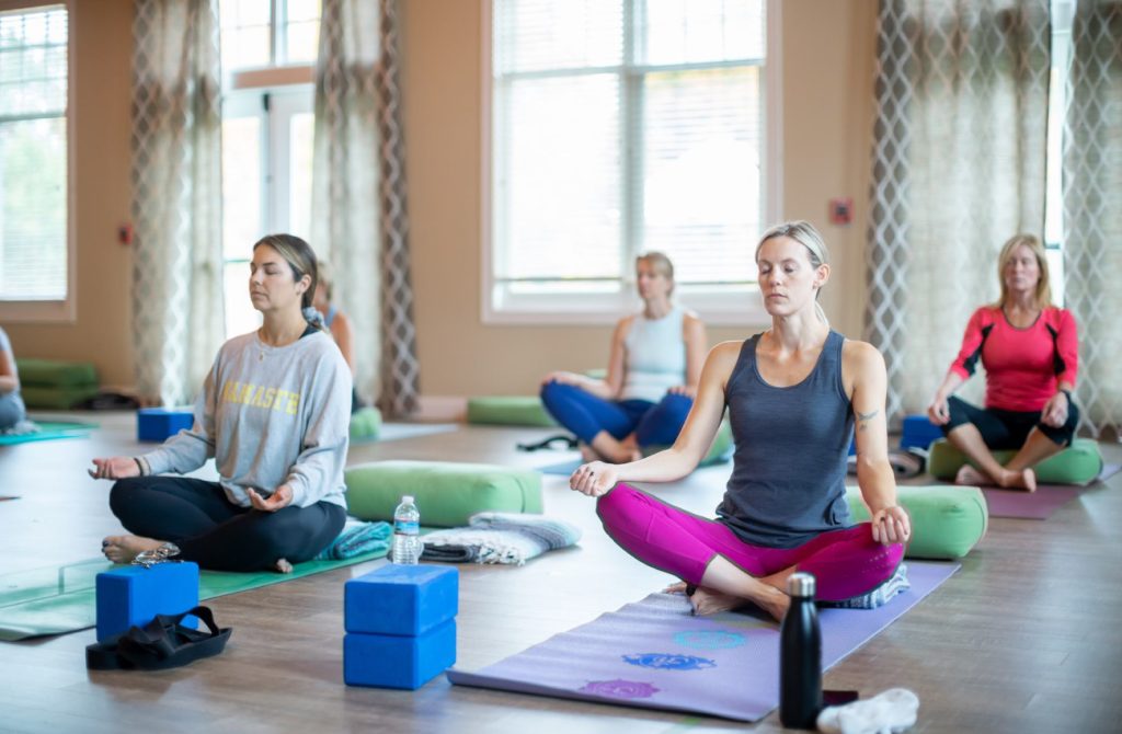 four women sitting on yoga mats indoors in a lotus pose with eyes closed
